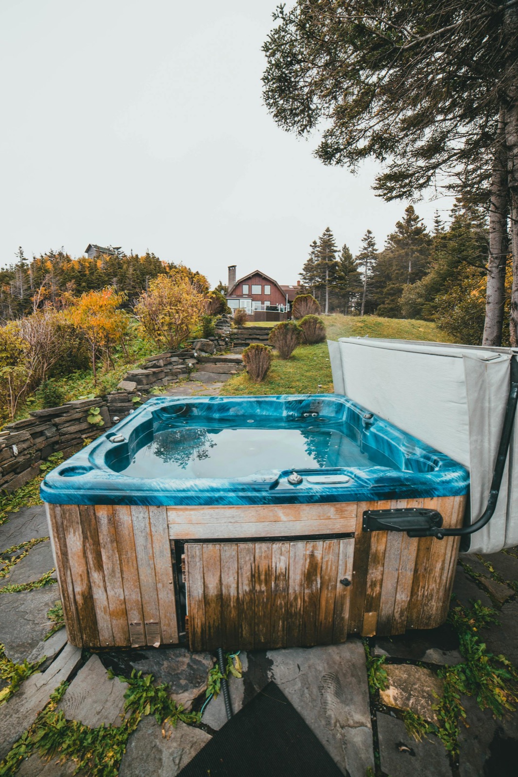 An outdoor spa on a stone patio with autumn foliage and an evergreen overhead — a small red cabin in the distance up the hill.