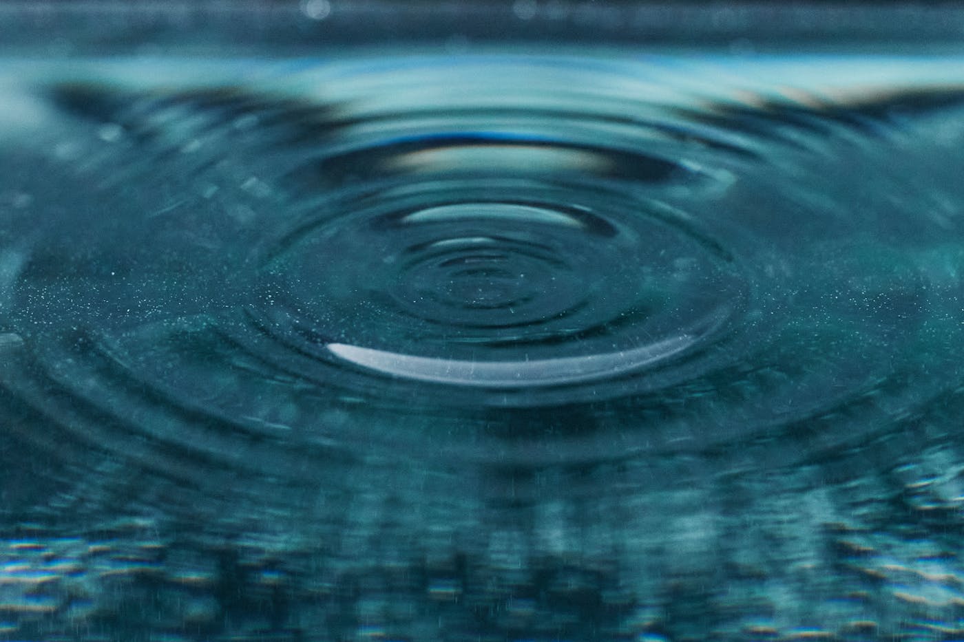 A single droplet rippling outward on a still teal pool surface, an extreme close-up.